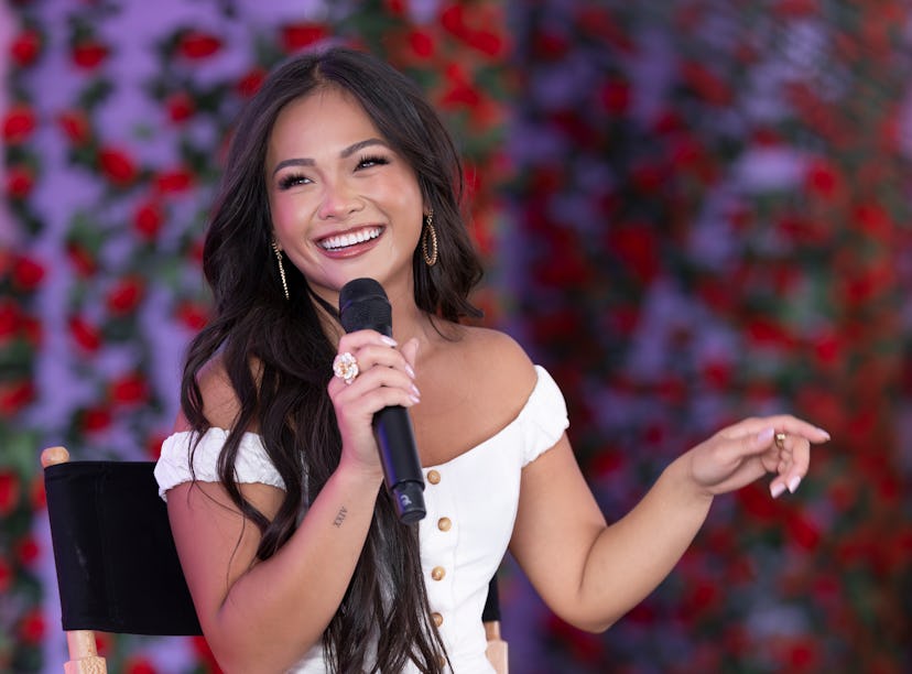 A joyful woman speaking into a microphone, sitting in front of a rose-covered background, wearing a ...